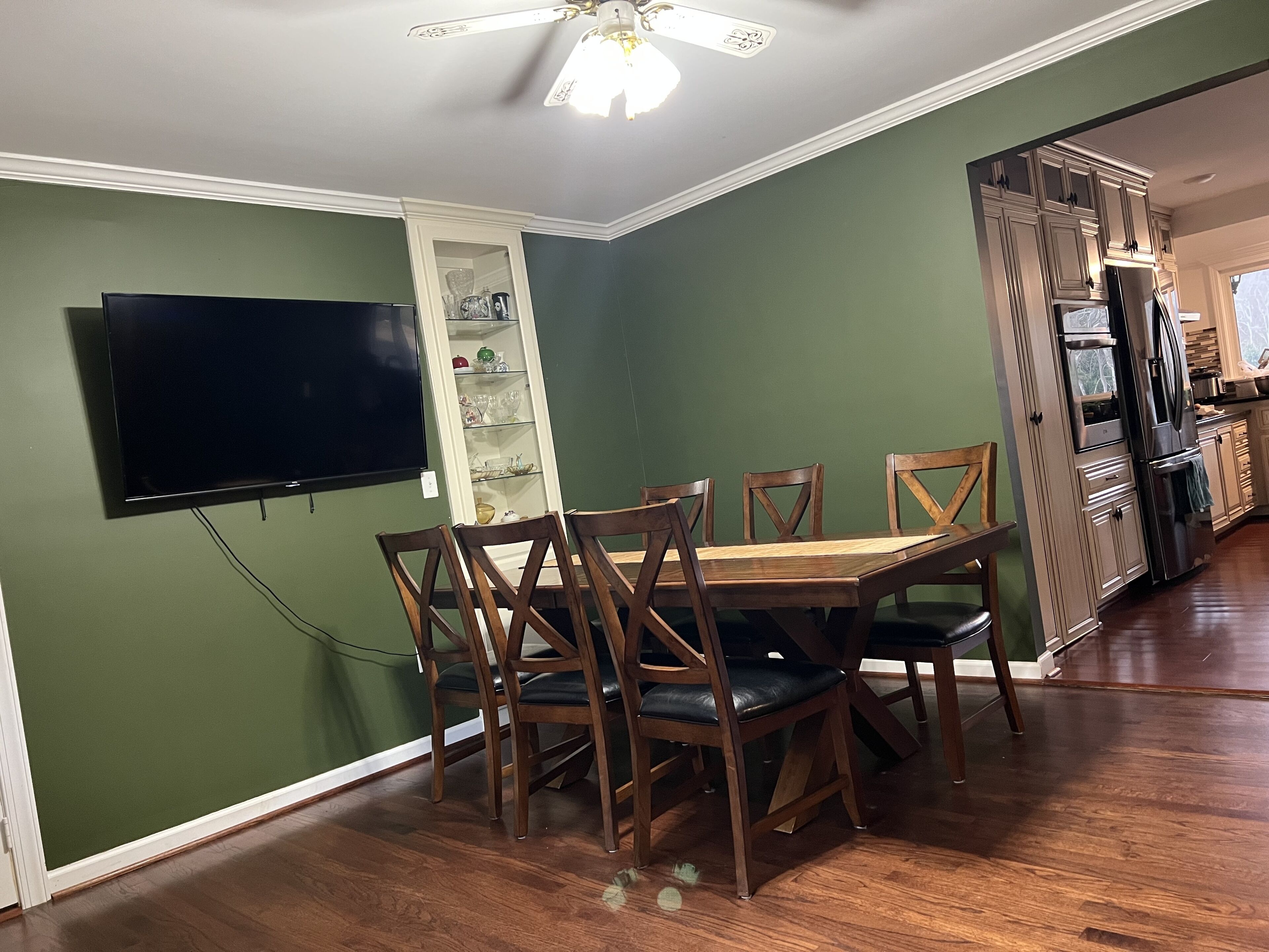 A dining area with a wooden table and chairs, a TV mounted on the wall, and a kitchen with cabinets and appliances.