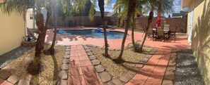 Un patio trasero con piscina, palmeras y un área de patio con sillas y una mesa.