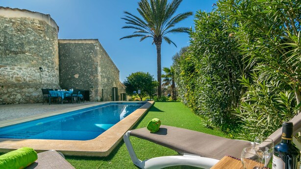 Una piscina con pared de piedra, una palmera y una mesa con una botella y un vaso.