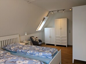 A bedroom with a sloped ceiling, a bed with floral bedding, a chair, a wardrobe, and a desk with a lamp.