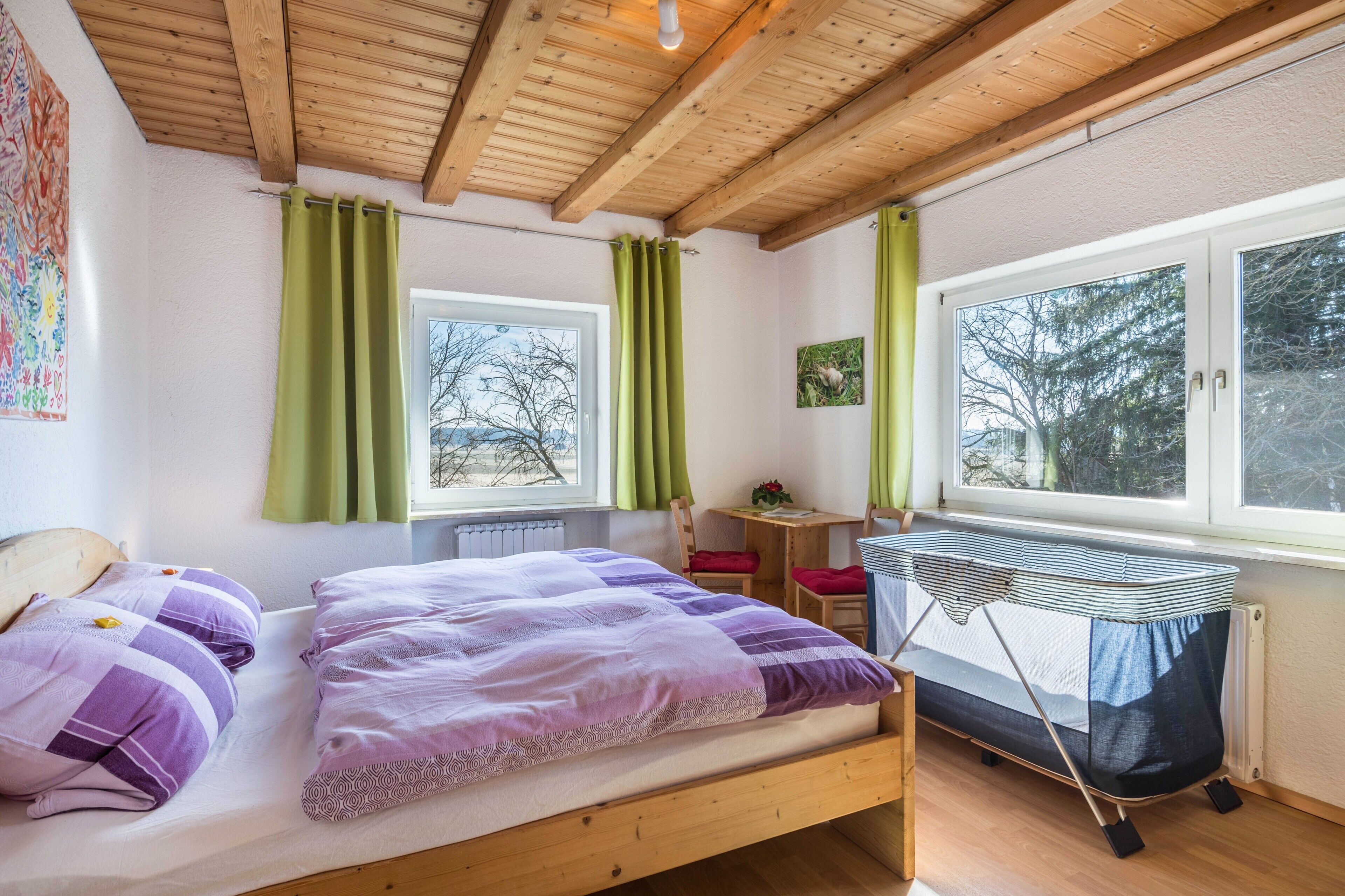 Un dormitorio con cama de madera, cortinas verdes, una ventana, un radiador y una mesita con una silla.