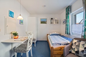 Una camera piccola con un letto, una scrivania, una sedia, un tavolino con un vaso e una finestra con tende.
