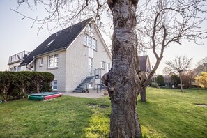 Una casa de dos pisos con una escalera visible que lleva al nivel superior, un trampolín verde en el jardín y un árbol en primer plano.
