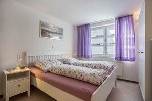 Un dormitorio con cama, mesita de noche, lámpara, ventana con cortinas moradas y un cuadro en la pared.