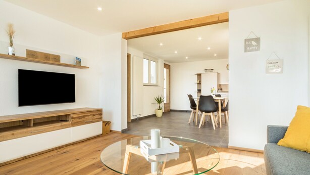 Uma sala de estar moderna com televisão de tela plana, móveis de madeira, uma mesa de centro de vidro e uma área de refeições ao fundo.