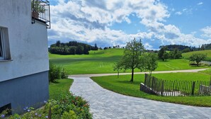Um caminho pavimentado leva a um edifício com varanda, rodeado por um jardim ajardinado com flores e vista para um campo verde e árvores.