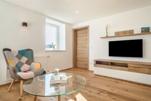 Ein modernes Wohnzimmer mit Glastisch, einem bunten Sessel, einem Flachbildfernseher, Holzboden und einem Fenster mit Blick.