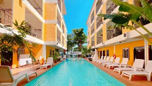 A swimming pool surrounded by white lounge chairs, with yellow buildings and greenery in the background. - Guyana Saikaew (Rayong)