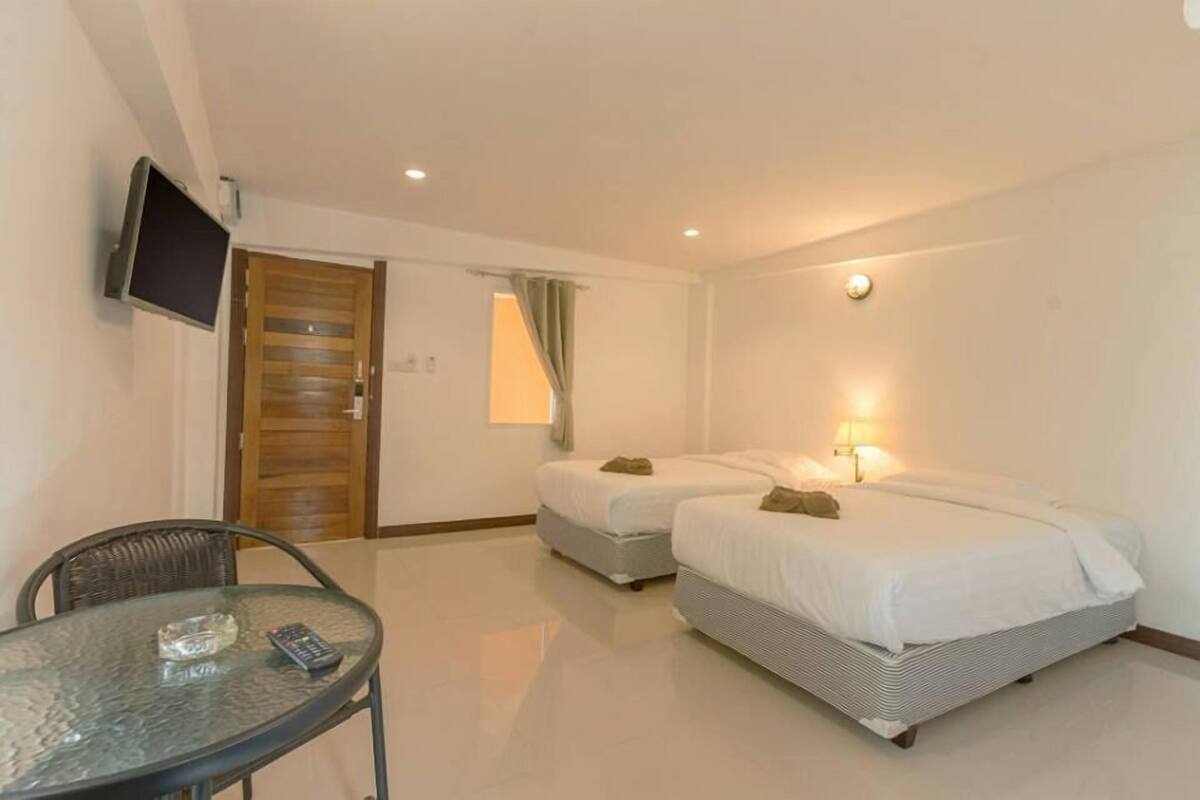 A hotel room with two single beds, a glass table with a remote control, a TV mounted on the wall, and a wooden door.