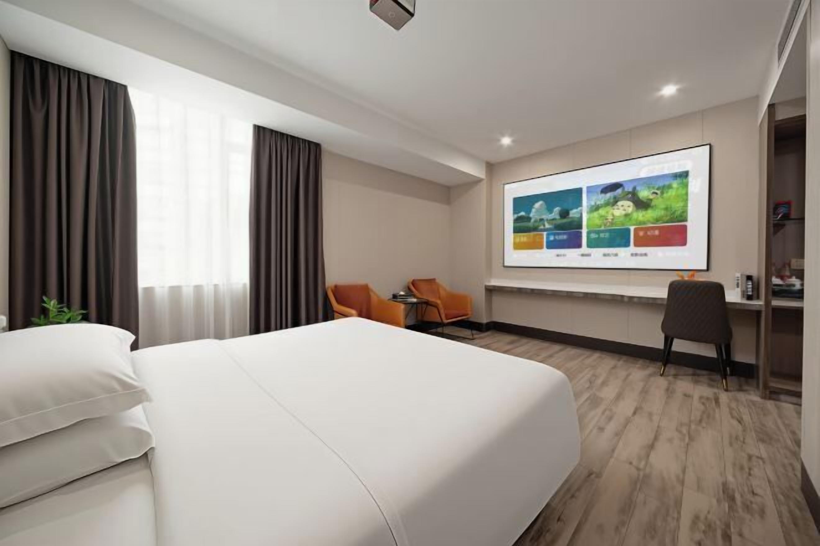 A hotel room with a bed, two chairs, a desk, and a large TV screen.