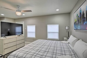 Un dormitorio con una cama, un televisor sobre una cómoda, un ventilador de techo y un cuadro de paisaje urbano.