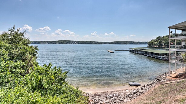 A lakeside view with a dock extending into the water, surrounded by greenery and a clear sky. - Water Views + Pool Access: Lake Ozark Escape! (Lake Ozark)
