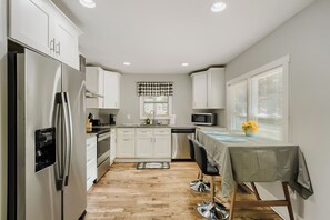 A modern kitchen with white cabinets, stainless steel appliances, a dining area with a table and chairs, and a window with blinds.