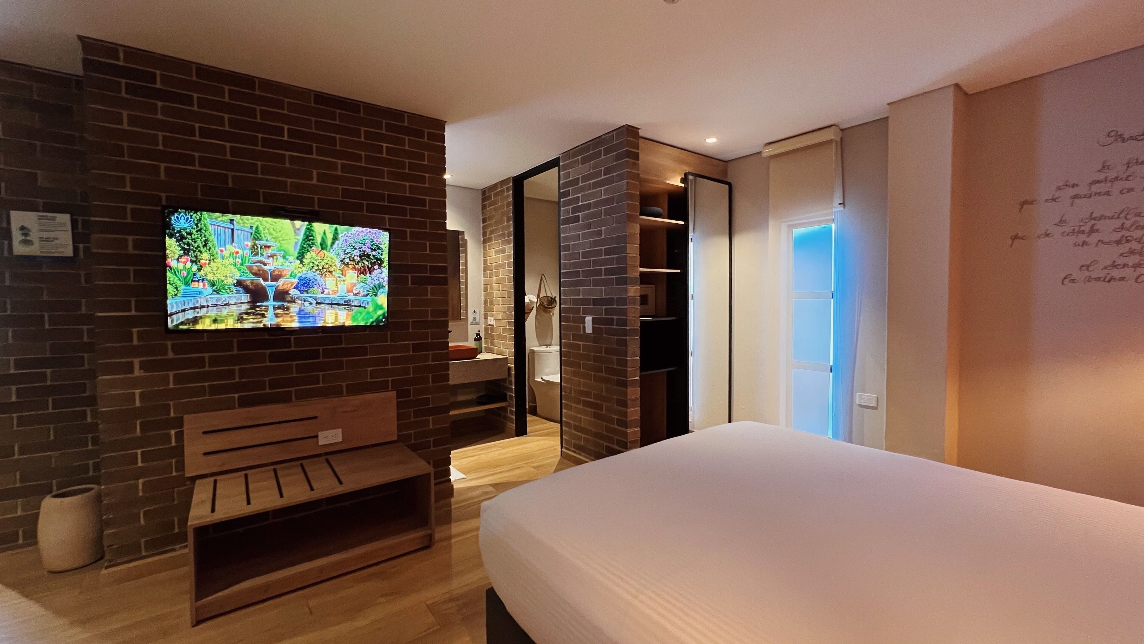 Quarto de hotel moderno com parede de tijolos, TV, um banco de madeira, cama e banheiro com chuveiro e vaso sanitário.