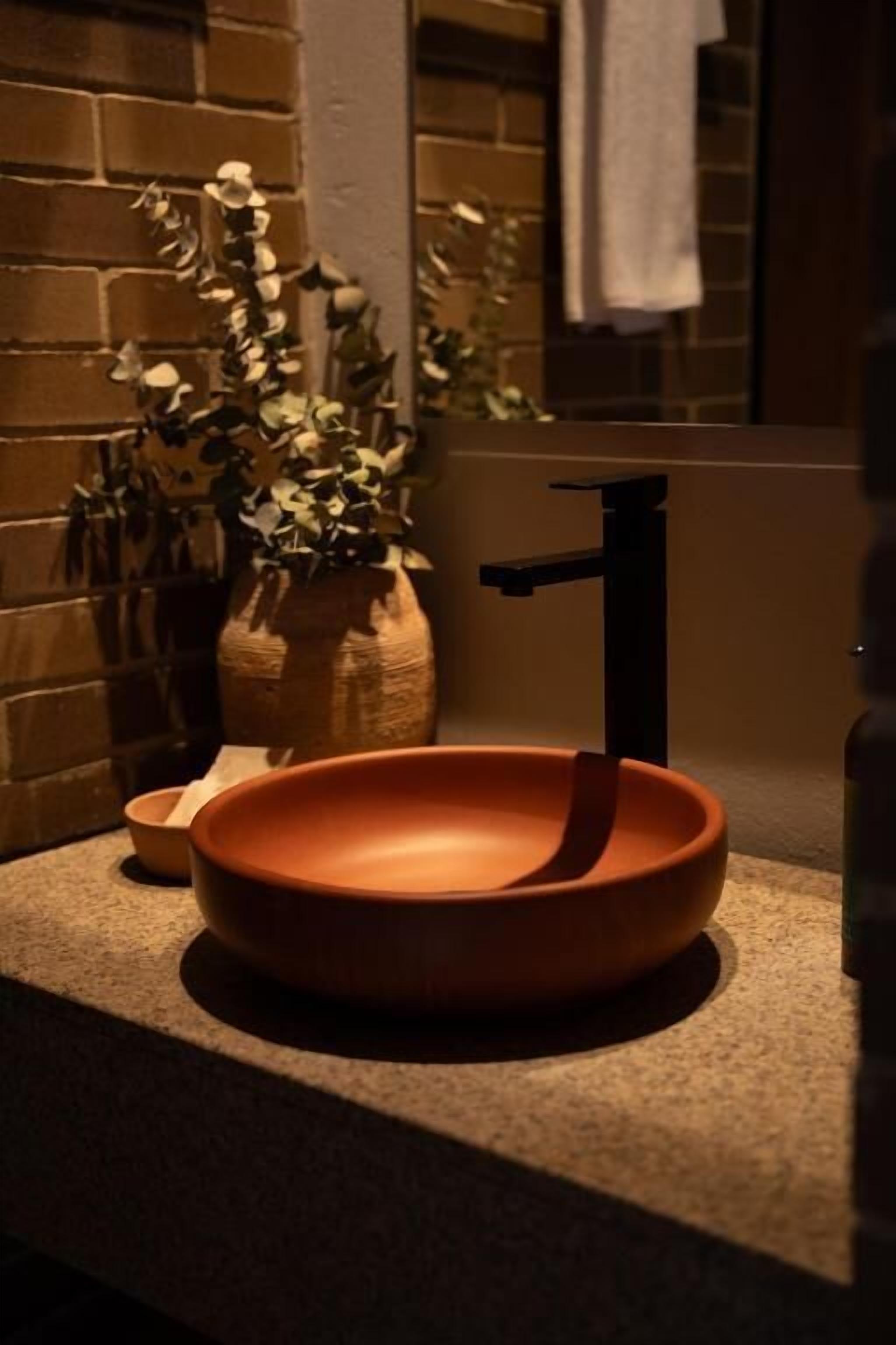 A bathroom with a round, terracotta sink, a black faucet, a potted plant, and a towel hanging on the wall.