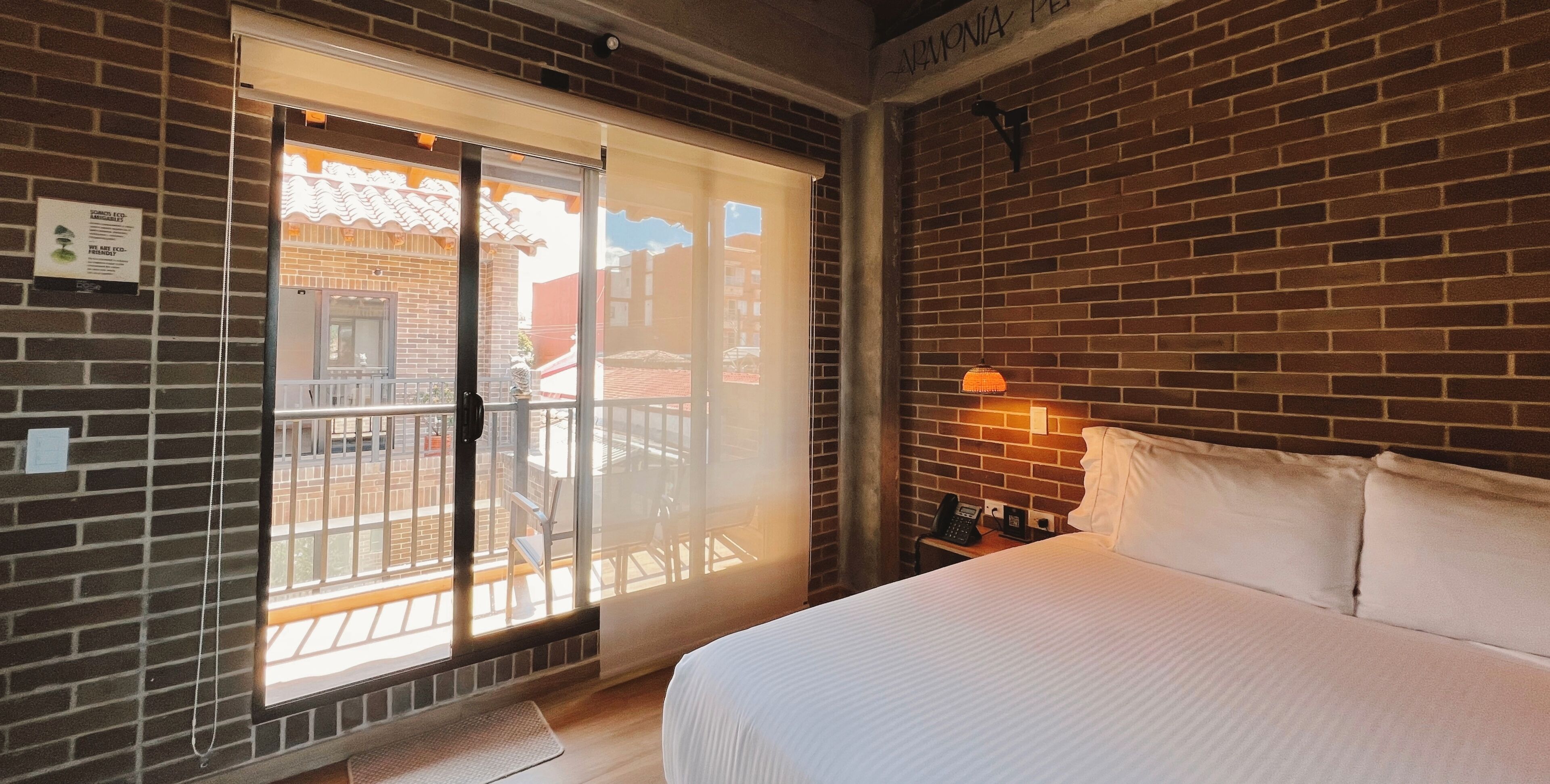 A hotel room with a brick wall, a bed with white bedding, a balcony with a view, and a phone on the nightstand.