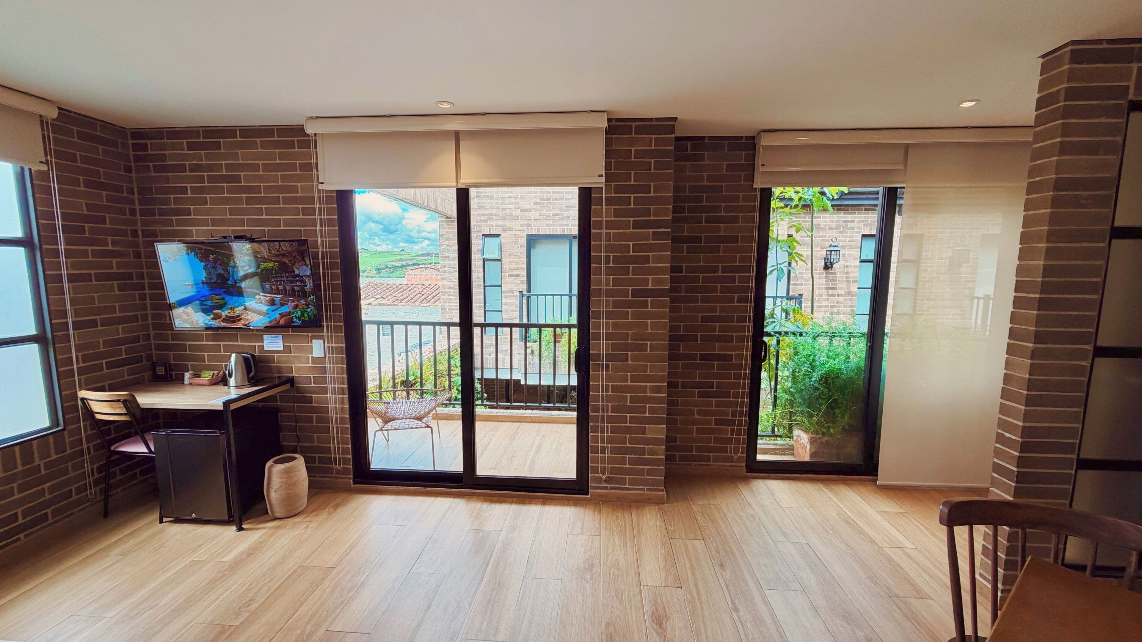 Quarto amplo com piso de madeira, uma escrivaninha com cadeira, TV fixada na parede e porta de vidro corria para uma varanda externa.