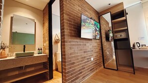 Un baño moderno con encimera de granito, un lavamanos, un espejo, un televisor montado en una pared de ladrillo y un refrigerador.