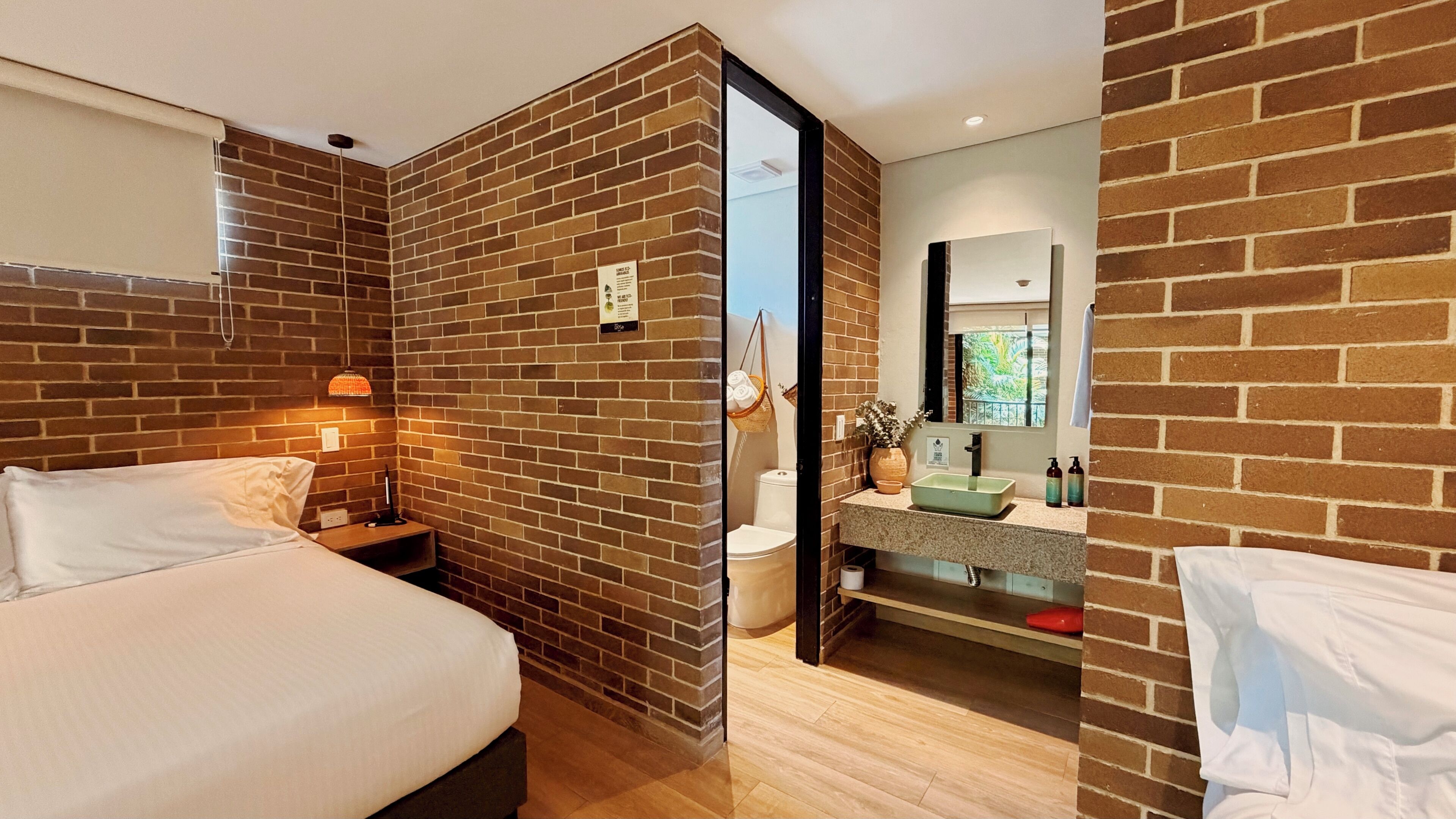 A hotel room with a brick wall, a bed, a nightstand, a bathroom with a sink and mirror, and a window with a view.