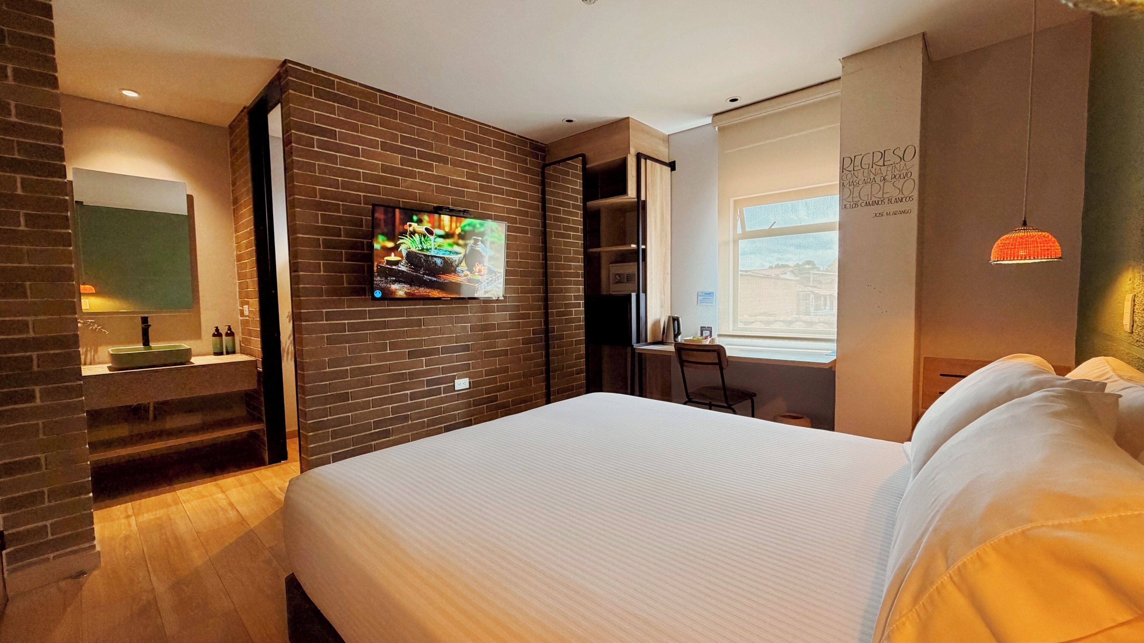 A modern hotel room with a large bed, a TV mounted on a brick wall, a desk with a chair, and a window with a view.