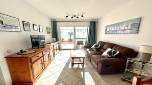 A living room with a brown sofa, a coffee table, a TV on a wooden cabinet, and a balcony with curtains. - WIFI+and next to the beach and in the centre (Costa Calma (incl. Urbanización Cañada del Río))