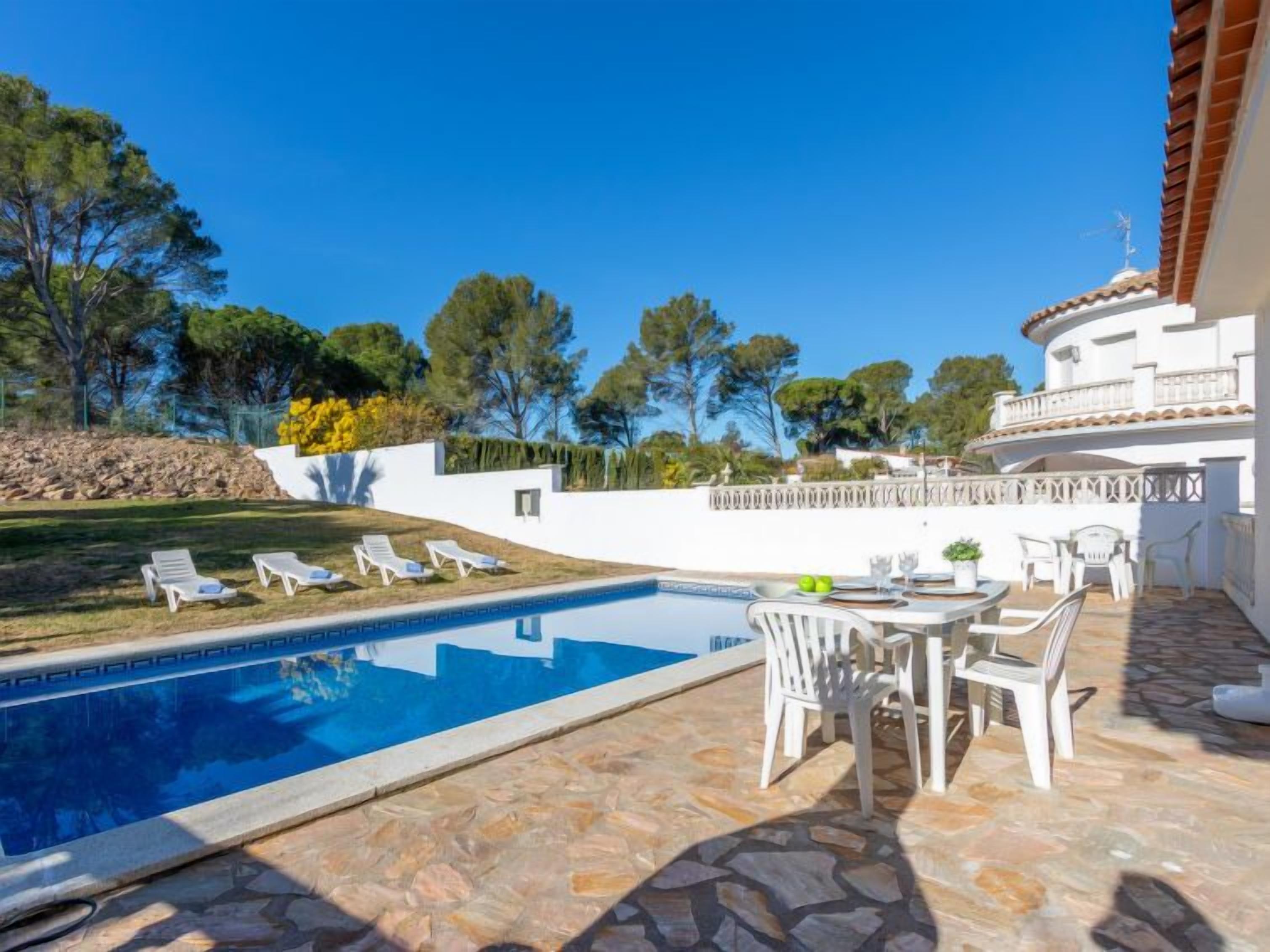 Un espace piscine avec des chaises longues, une table dressée pour les repas et un bâtiment blanc doté d’un toit en tuiles.