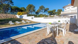 A pool area with lounge chairs, a table set for dining, and a white building with a tiled roof. - Villa Marc by Interhome (L'Escala)