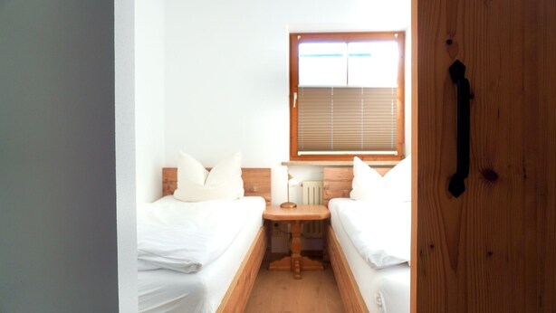 A small, bright room with two single beds, a wooden nightstand, a window with blinds, and a door with a handle. - Three-room vacation apartment 2, approx. 63 sqm with balcony and wonderful view (Reit im Winkl)