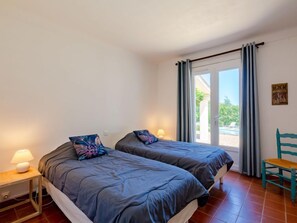 Quarto com duas camas, uma cadeira, um abajur e vista para o exterior.