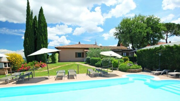 Una piscina con sillones de descanso y sombrillas, rodeada por un jardín cuidado y una casa con techo de tejas.
