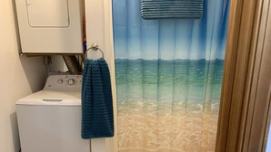 A laundry area with a washing machine, a drying rack, and a curtain depicting a beach scene. - View Peaceful Pond while lounging in this open studio apartment! (Warsaw)