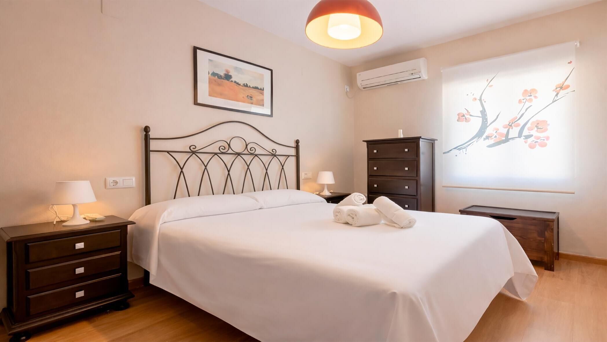 Una cama bien hecha con ropa blanca, una mesita de noche de madera oscura con una lámpara, un aparador con cajones y un aire acondicionado instalado en la pared.