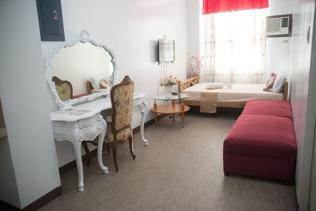 A bedroom with a bed, a desk, a chair, a mirror, and a small table.