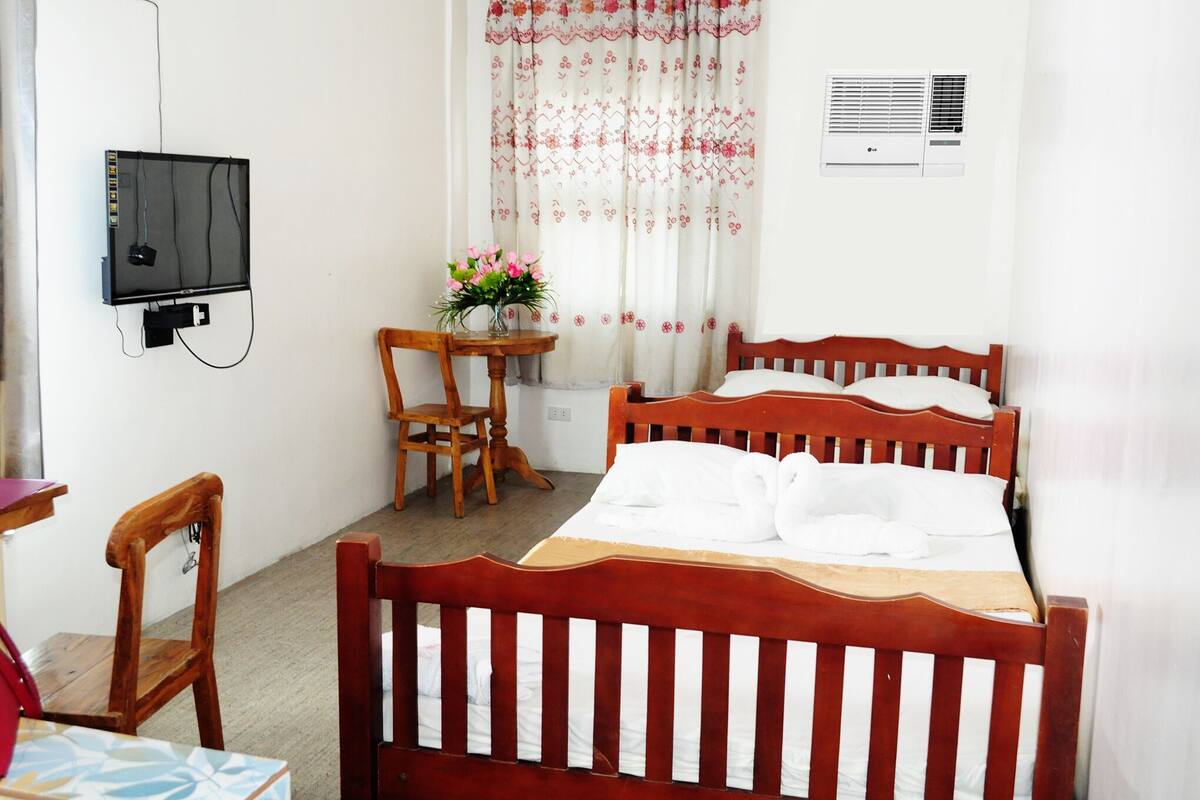 A bedroom with a wooden bed, a desk with a chair, a television mounted on the wall, and a window with floral curtains.