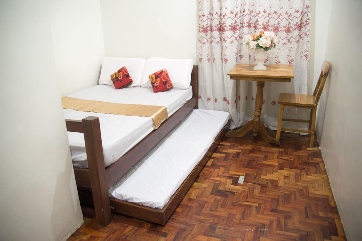 A small room with a bed, a wooden chair, a small table with a vase of flowers, and a herringbone-patterned floor.
