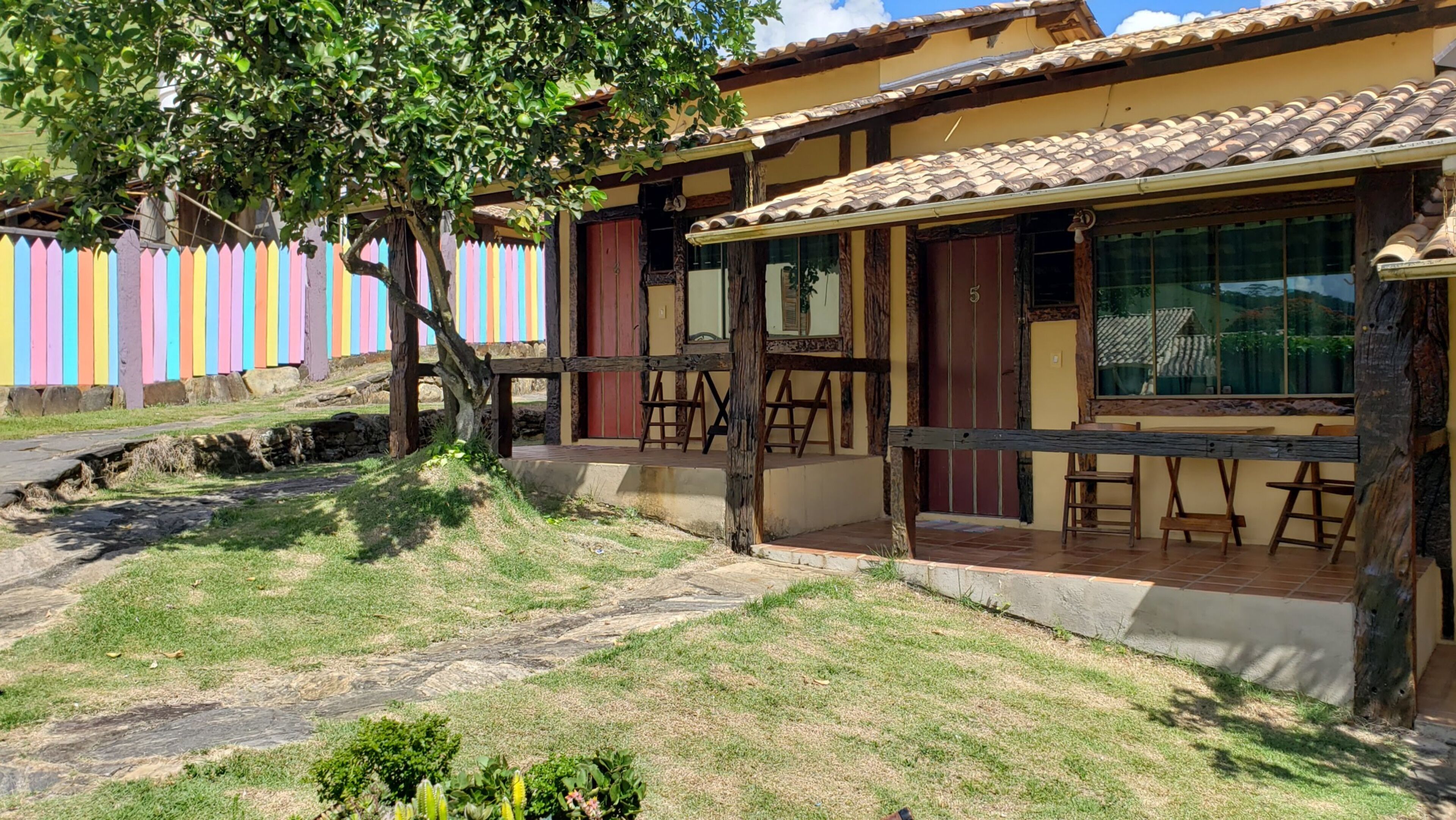 A two-story building with a tiled roof, a porch with wooden chairs, and a colorful fence in the background.