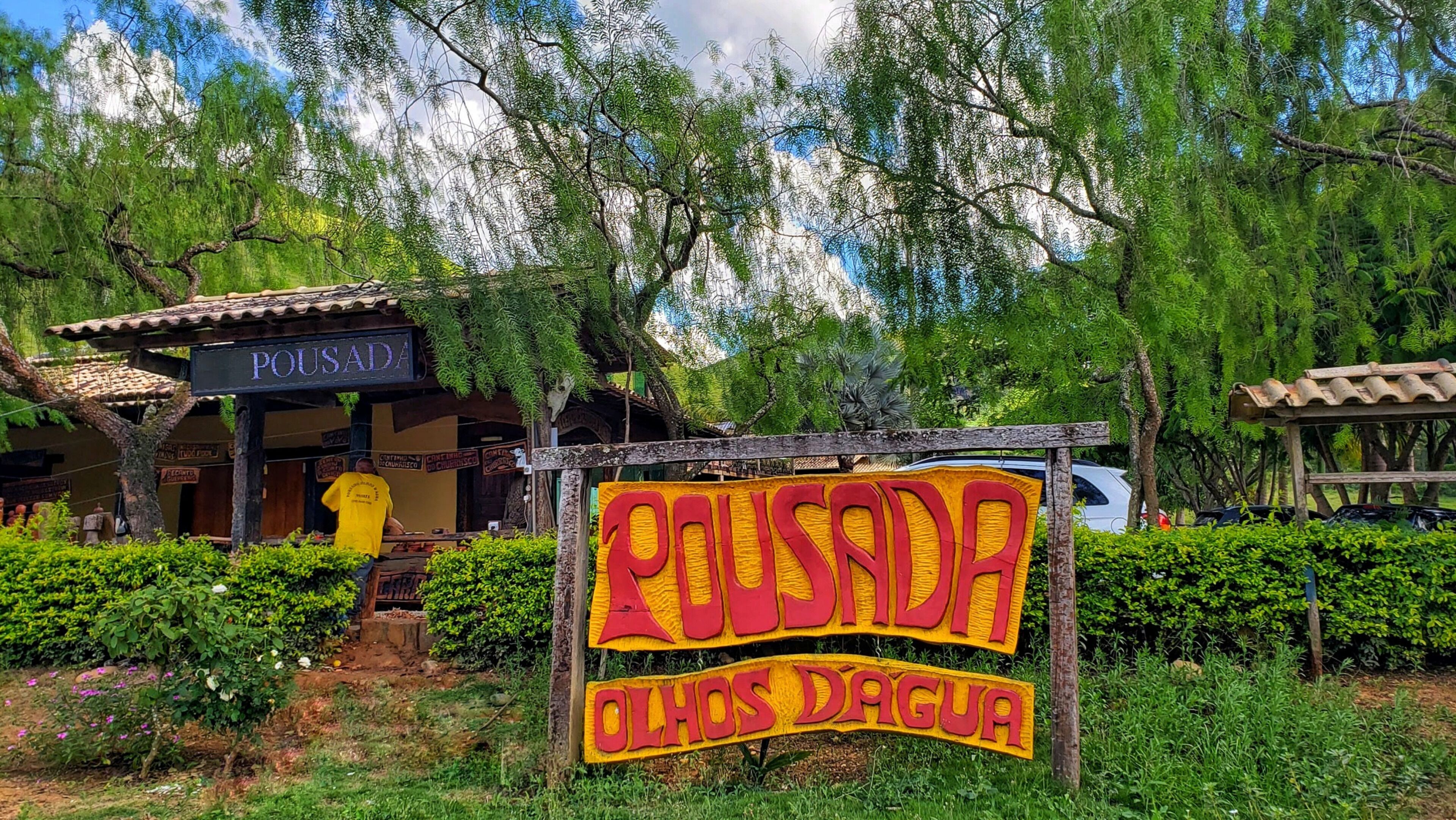 Un bâtiment rustique, sur lequel on peut lire l’inscription « POSADA », et une autre inscription indiquant « PUSADA OLHOS D’ARGUA », le tout dans un environnement luxuriant et verdoyant.