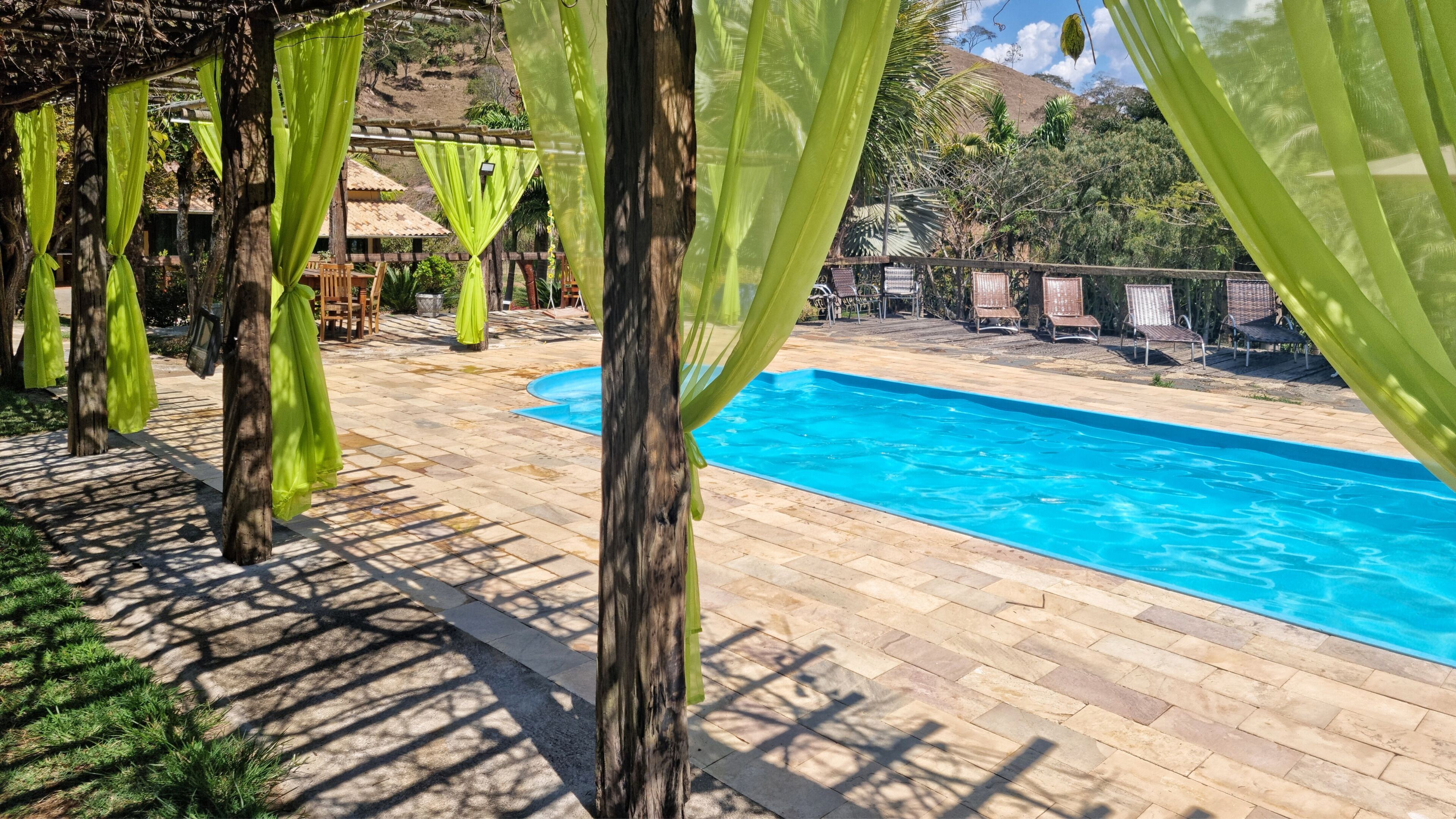 Una piscina con deck de baldosas, rodeada de toldos verdes y sillas de madera.