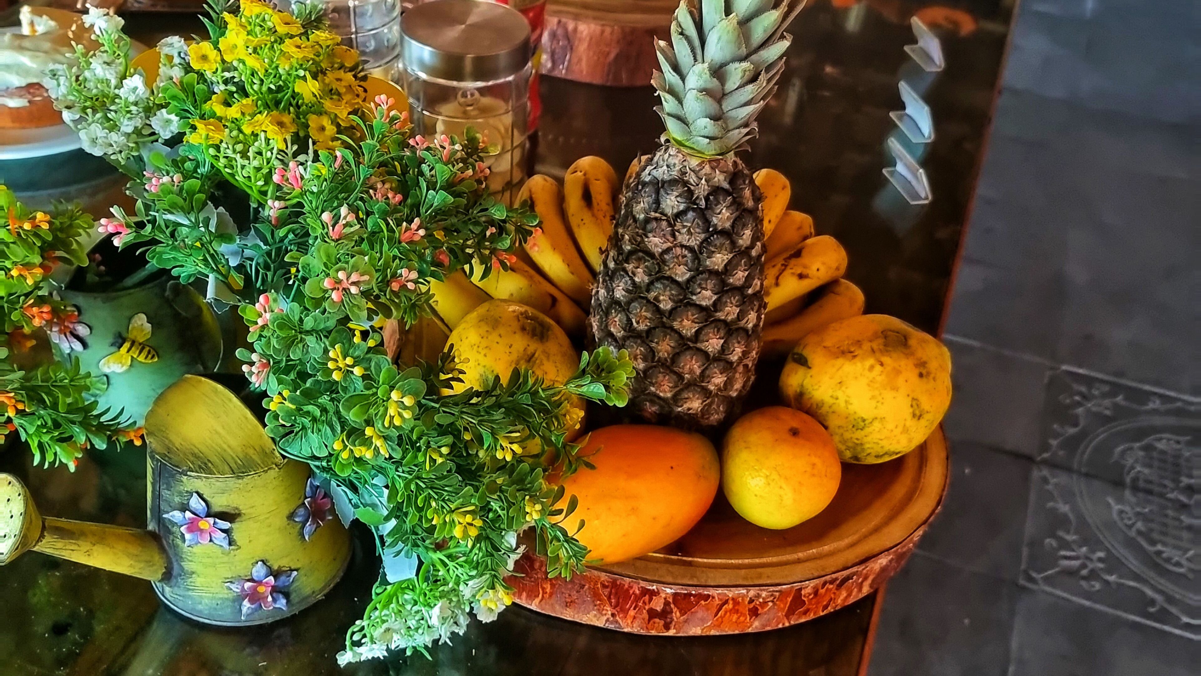 Una mesa con un frutero que contiene plátanos, una piña y naranjas.