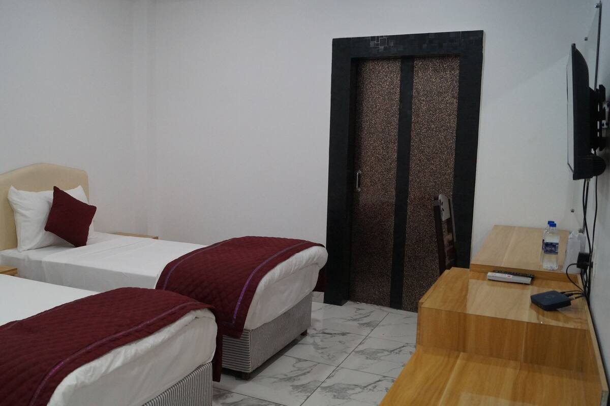 A hotel room with two beds, a wooden nightstand, a TV, and a door with a textured panel.