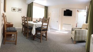A dining area with a table and chairs, a radiator, a television on the wall, and a door leading to another room. - Lategillan Cottage -  a cottage that sleeps 8 guests  in 3 bedrooms (Tweedsmuir)