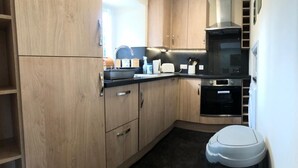 A compact kitchen with wooden cabinets, a black countertop, a built-in oven, and a white trash bin. - Lategillan Cottage -  a cottage that sleeps 8 guests  in 3 bedrooms (Tweedsmuir)