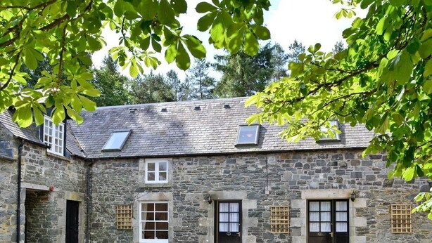 A stone building with a courtyard, featuring a central door, two windows with shutters, and a small window with a grid pattern. - Lategillan Cottage -  a cottage that sleeps 8 guests  in 3 bedrooms (Tweedsmuir)