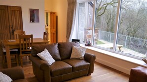 A cozy living room with a brown leather sofa, matching armchairs, and a dining table. A large window offers a view of the outdoors.