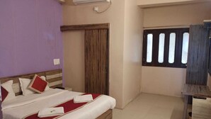 A hotel room with a bed, a wooden headboard, a window with blinds, and a small table. - Hotel Always Welcome (Abu Road)