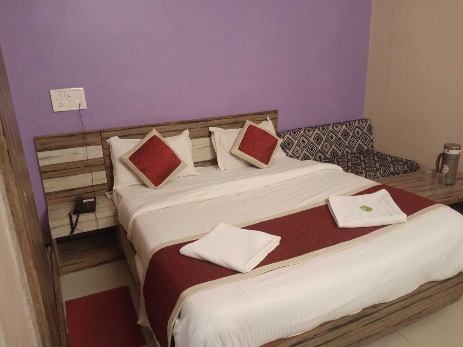 Una cama bien hecha con cabecera de madera, almohadas y colcha rojas y blancas. Hay una mesita de noche con una lámpara y una taza.