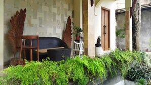 A covered outdoor seating area with a wooden chair and a wicker sofa, surrounded by lush green plants and a stone wall. - Villa Kania Parangtritis (Sewon)