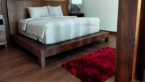 A bed with a wooden frame and white bedding, a red rug on the floor, and a nightstand with a lamp. - Villa Kania Parangtritis (Sewon)