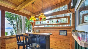 Een houten interieur met een bar, twee stoelen, een bord met Heineken erop, en uitzicht op bomen en water.