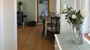 A wooden-floored dining area with a table and chairs, a vase with flowers, and a kitchen visible through an open doorway. - Cozy Scandinavian-style vacation home between the Schlei and the Baltic Sea (Rieseby)